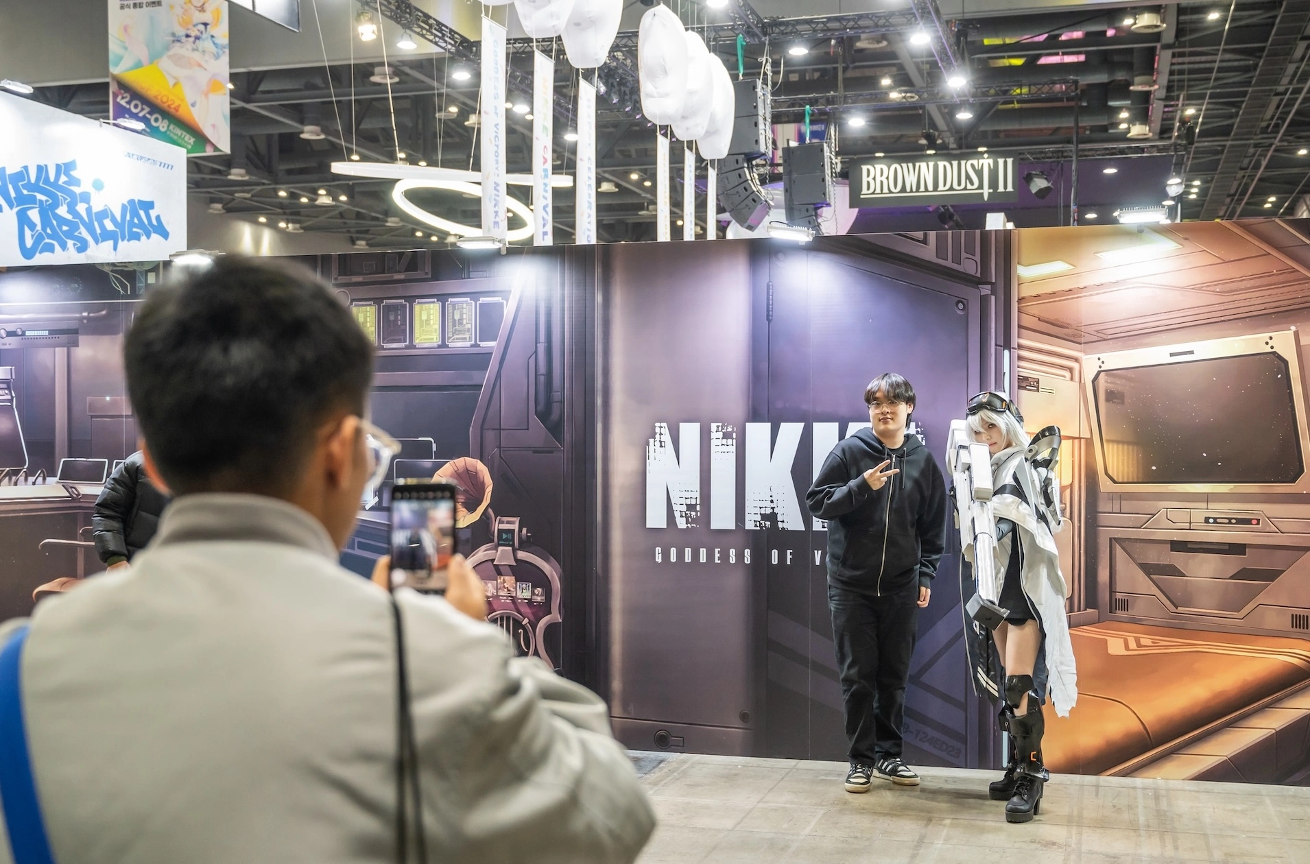 Visitor taking a photo with a NIKKE cosplayer in the AGF exhibition hall, NIKKE and Brown Dust 2 banners overhead