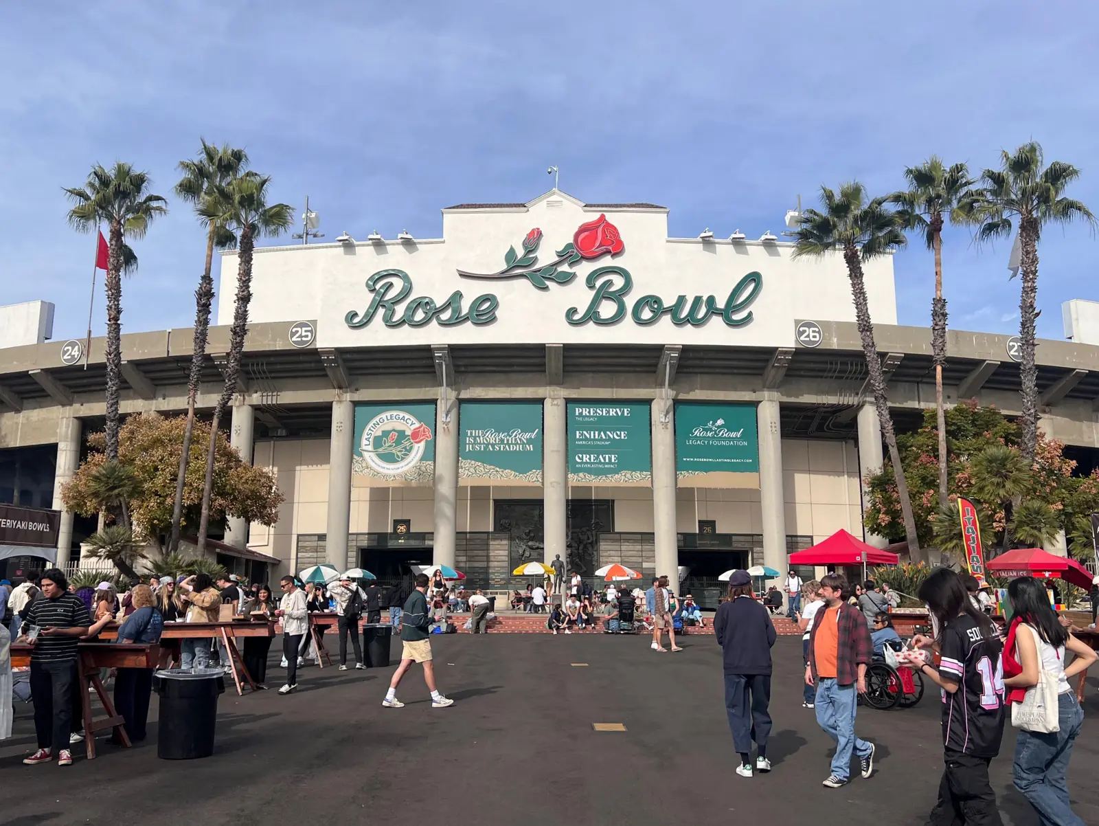 Main entrance of the Rose Bowl Stadium in Pasadena, California, with the iconic script signage and rose emblem above Gates 24–26, framed by palm trees and visitors gathering on the plaza